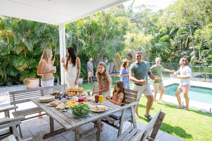 A group of people enjoy a backyard gathering by a pool during a sunny July campaign. Some chat near the patio table set with food, while others play and converse by the pool, all surrounded by lush greenery.