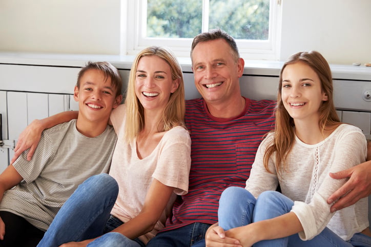 A smiling family of four, including two adults and two children, sits closely together indoors near a window, posing for a cheerful group photo as part of the META campaign.