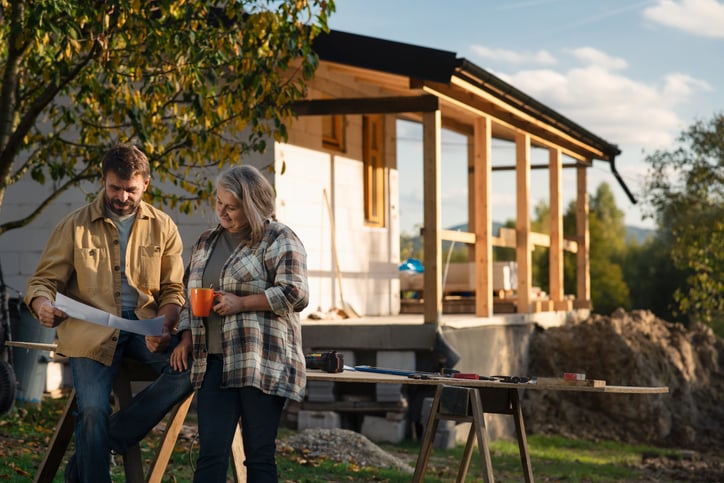 A man and a woman stand outside a house under construction, looking at blueprints—perhaps planning renovations or even considering refinancing for renovations. The woman holds an orange mug, with tools and materials spread out before them amid lush greenery.