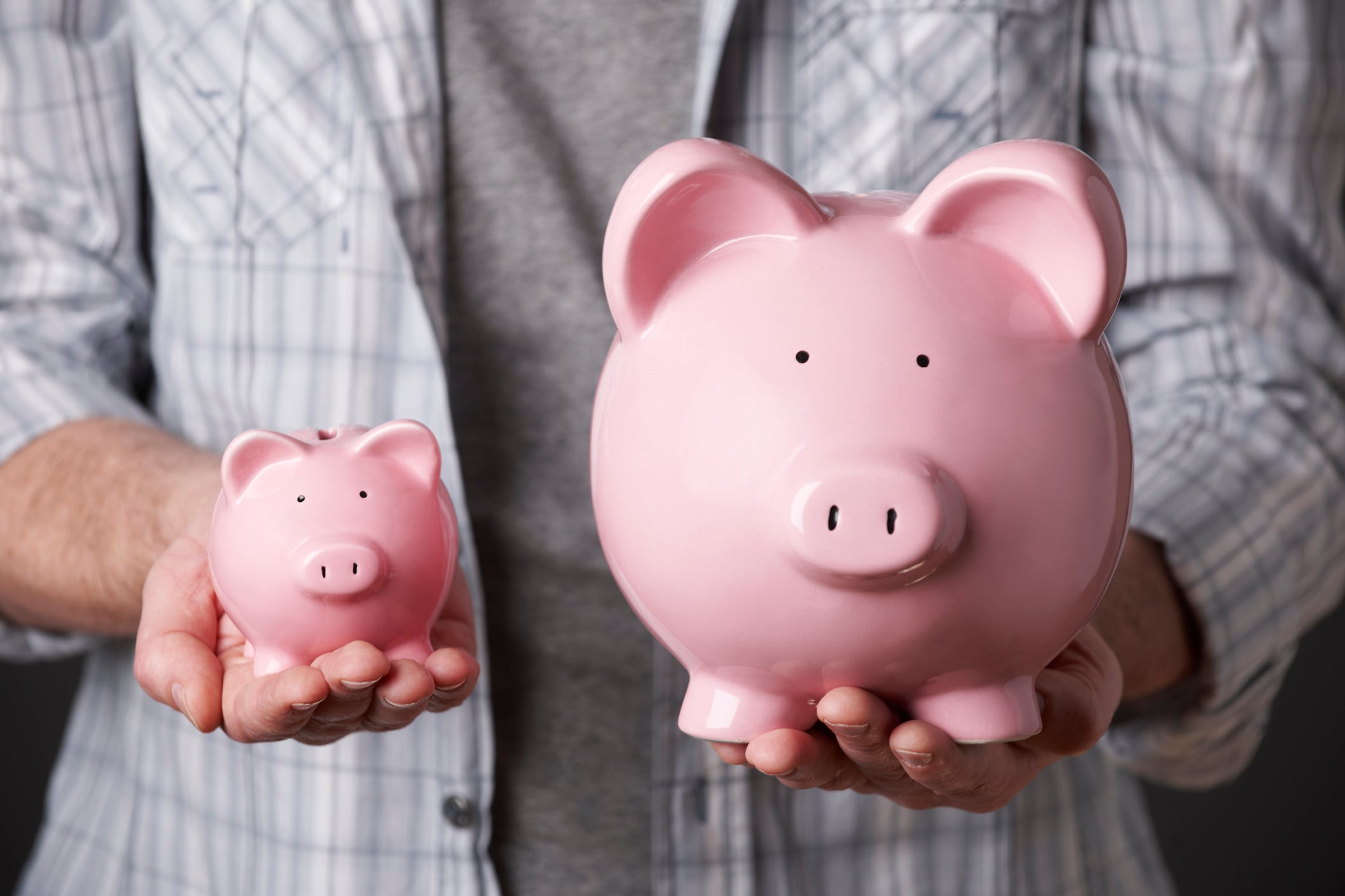 A person holding a small pink piggy bank in one hand and a large pink piggy bank in the other, both in front of their torso, wearing a plaid shirt—symbolizing the growth of retirement savings or a KiwiSaver review.
