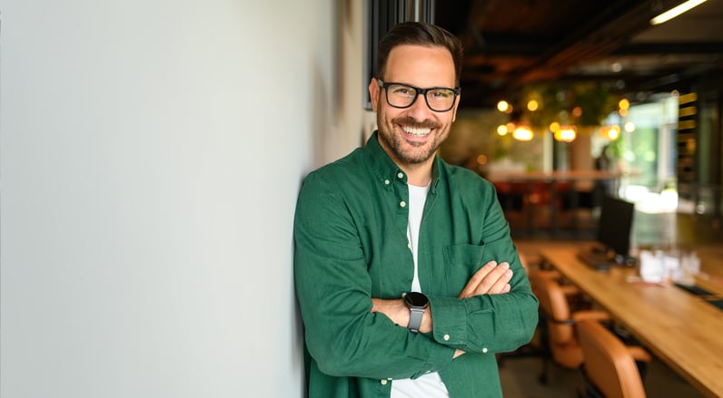 A smiling self-employed man with glasses and a green shirt stands with his arms crossed, leaning against a wall in a modern office with warm lighting and wooden desks in the background.