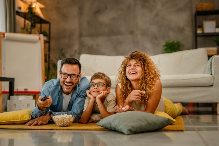 A cheerful family of three—a man, a woman, and a young boy—lie on the floor in front of a sofa, laughing and watching TV together with a bowl of popcorn nearby, celebrating their recent mortgage cashback win.