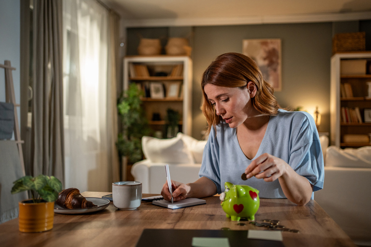 A woman sits at a table in a cozy living room, writing in a notebook while holding coins and using a green piggy bank—perhaps weighing cost vs value as she reviews her insurance coverage. A cup of coffee, a croissant, and some coins are also on the table.