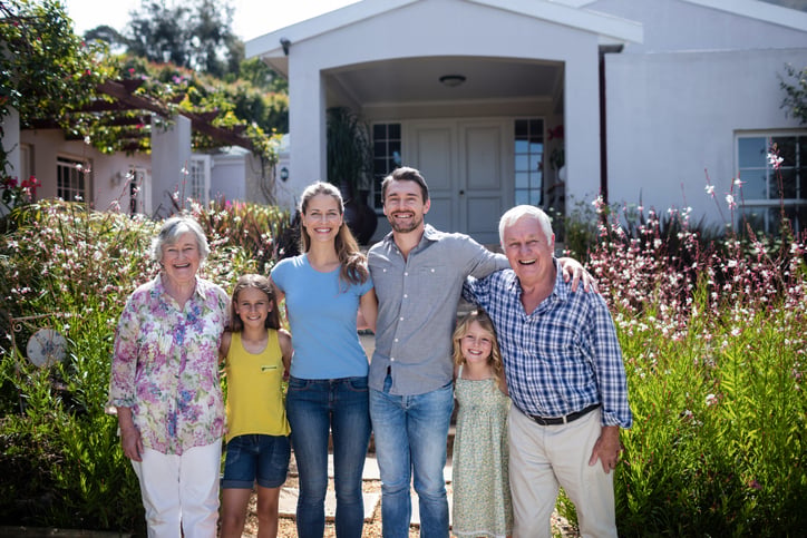 Three adults and two children stand smiling together in front of a white house with a garden, enjoying a sunny day—an ideal moment to reflect on KiwiSaver by age and securing retirement savings for every generation.