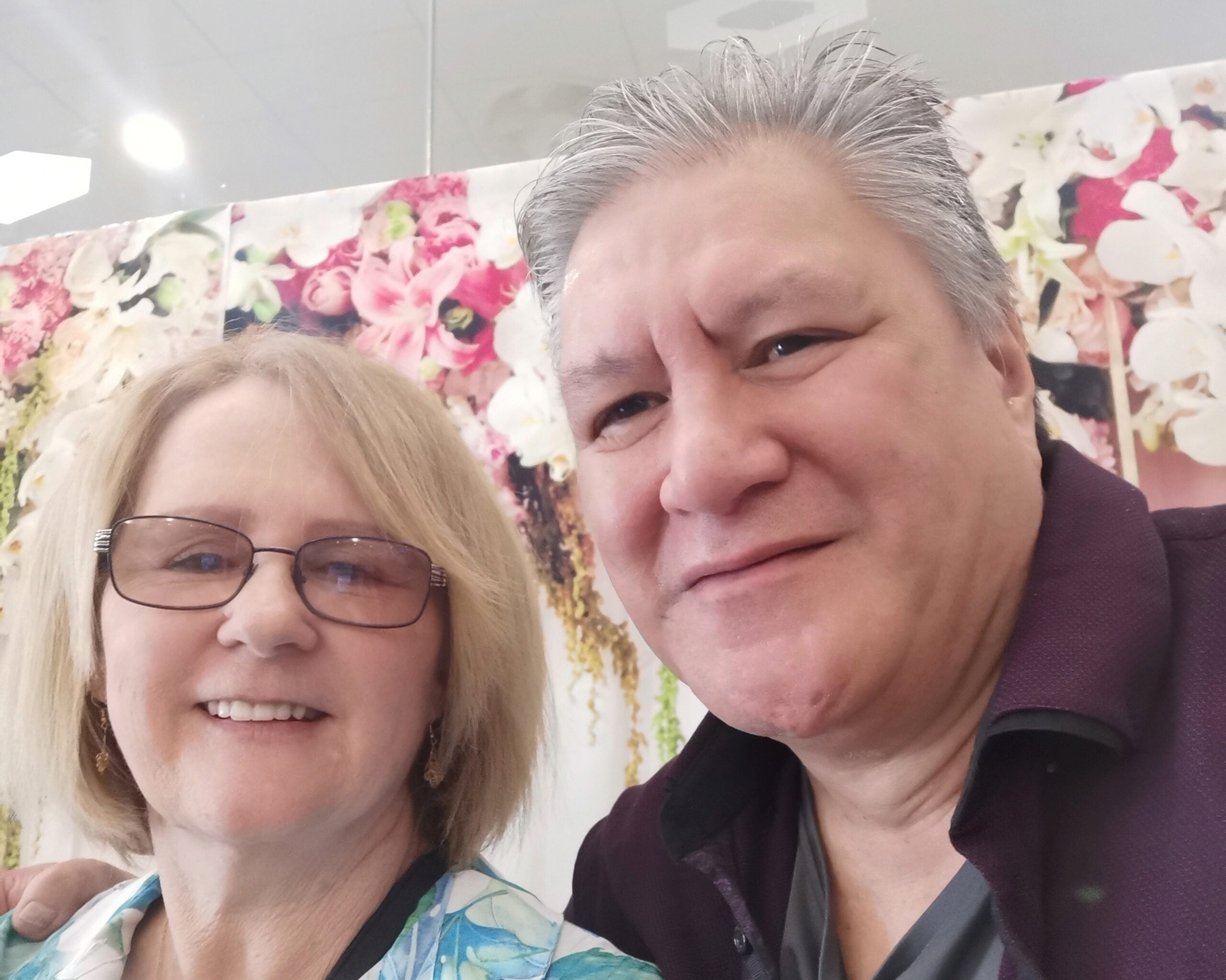 A smiling man and woman pose for a selfie in front of a floral backdrop with pink and white flowers, capturing the joy of a true winner moment. The woman has short blonde hair and glasses; the man has short gray hair and wears a dark shirt.