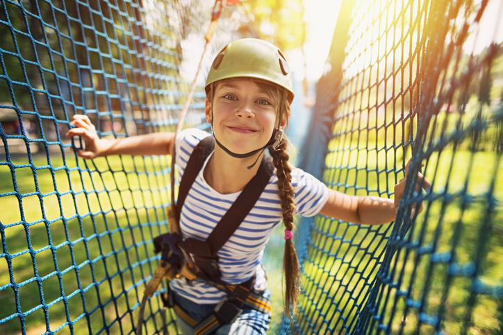 Smiling young girl wearing a helmet and safety harness stands between blue safety netting on a ropes course, lit by bright sunlight in an outdoor adventure park—a real life moment of fun and excitement.