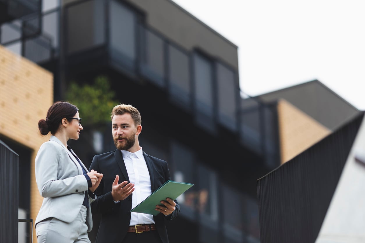 Two business professionals in suits stand outside a modern building, discussing interest-only loans and investment strategies. One holds a green folder as they engage in conversation against a backdrop of contemporary architecture.
