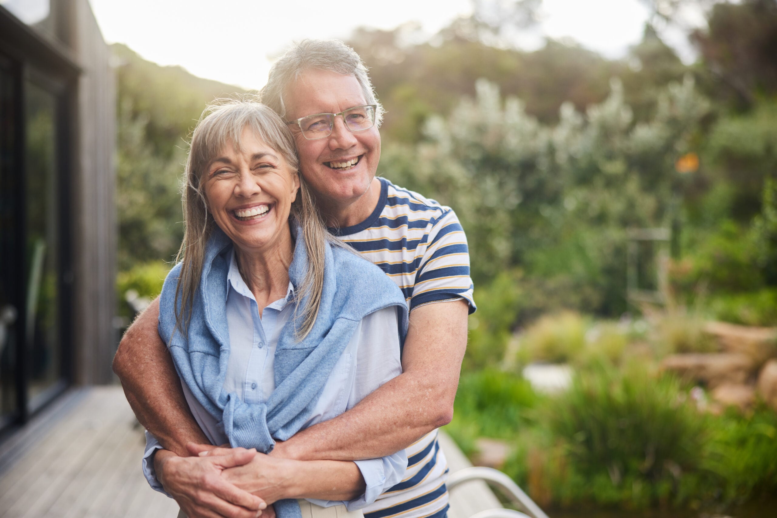 Smiling older couple, both over 65, standing outside with the man hugging the woman from behind. They appear happy and relaxed, enjoying a sunny day in a lush green garden or backyard.