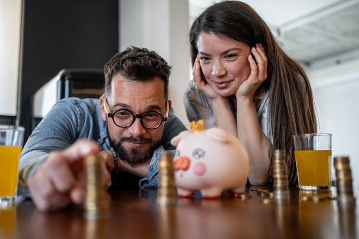 A man and woman smile while stacking coins on a table beside a piggy bank, glasses of orange juice, and Win Mortgage papers, suggesting they are saving money together for their home loan.