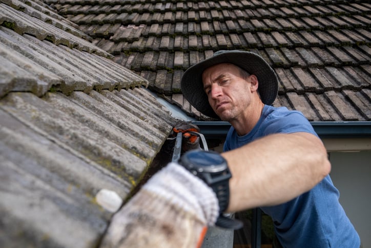 A man wearing a wide-brimmed hat and blue shirt cleans the gutter of a tiled roof with a gloved hand, showing preparedness as he focuses on his task before summer storms arrive.
