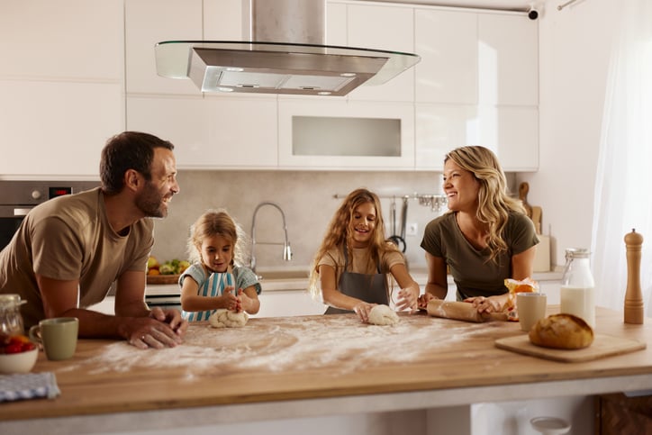 A family of four, including two children and two adults, smiles and enjoys making dough together at a kitchen island—just like the teamwork needed in mortgage planning for a successful future.
