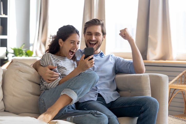 A joyful couple, Matt and Kristen, sit on a couch, celebrating and smiling while looking at a smartphone. The woman raises her arm in excitement as the man clenches his fist—both radiate congratulations and enthusiastic happiness.