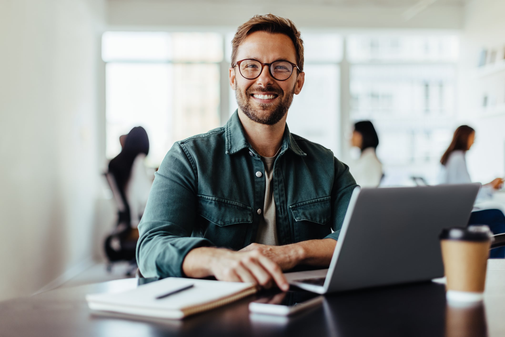 A smiling man with glasses sits at a desk with a laptop, notebook, and coffee cup, planning his career. He is wearing a green jacket over a white shirt. The bright office background shows others working at different life stages.
