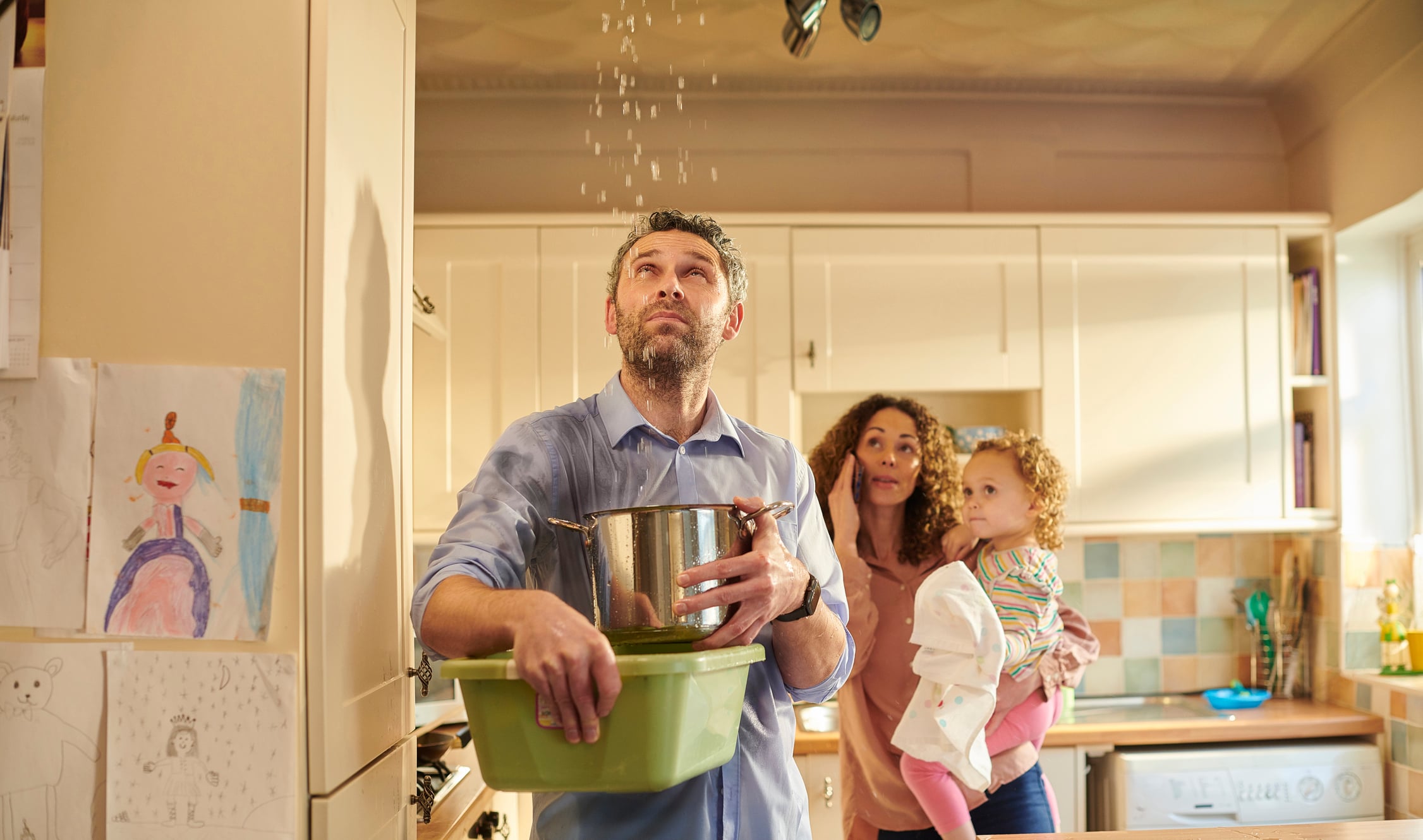 A man stands in a kitchen catching leaking water from the ceiling with a pot and a tub, while a woman holding a small child watches with concern in the background—reminding them of the importance of insurance and support during unexpected emergencies.