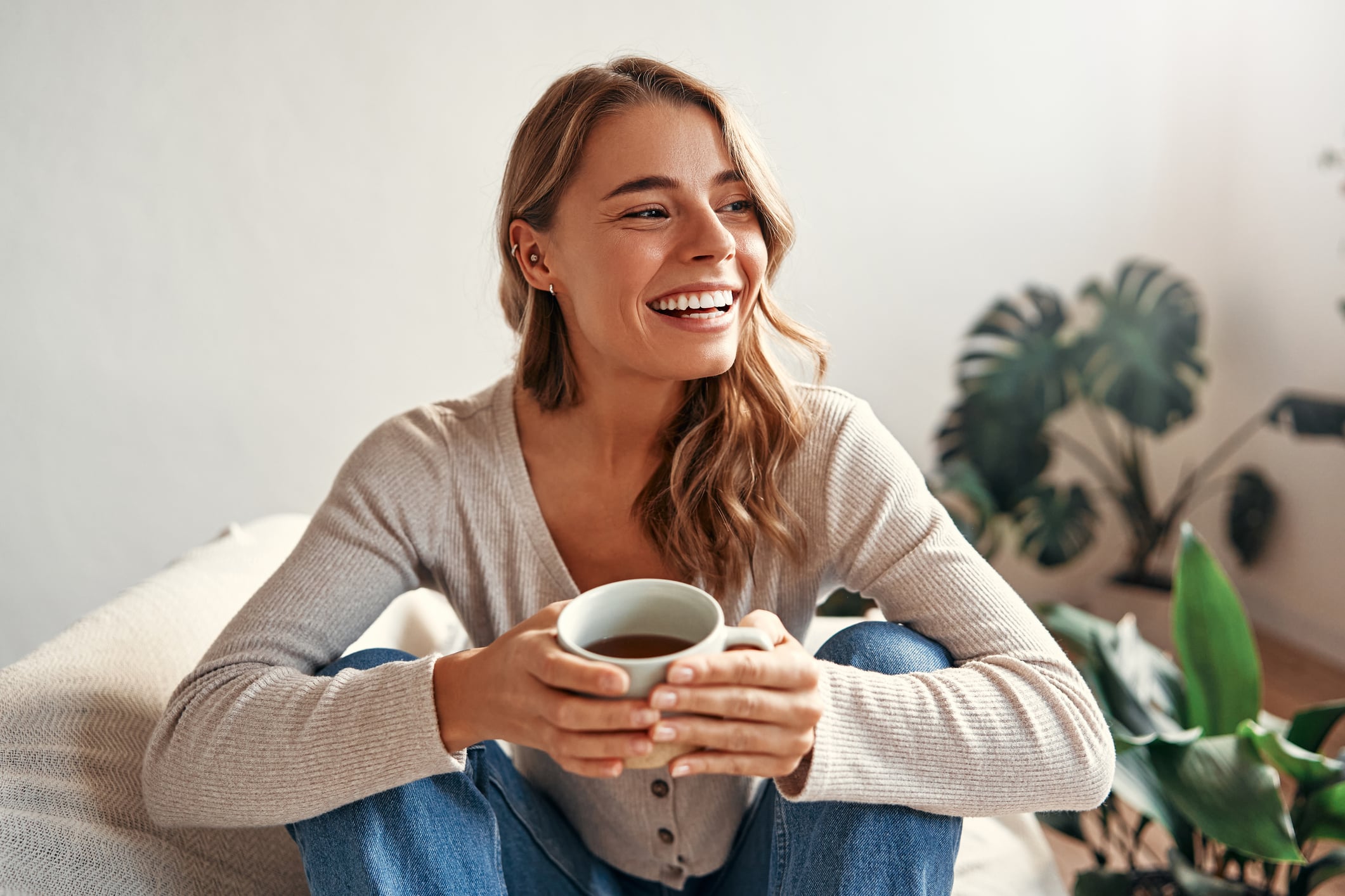A young woman with long light brown hair sits cross-legged indoors, smiling and holding a mug. She wears a light sweater and jeans, with green houseplants in the background and soft natural light as she checks her loan eligibility online.