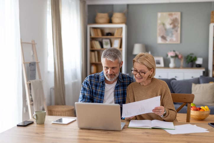 An older couple sits at a wooden table, smiling as they review documents and explore personal insurance options on a laptop in a cozy living room with bookshelves and home decor in the background.