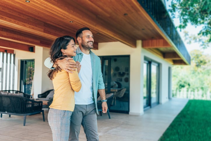 Happy couple embracing outside a large modern home - illustrating the decision to upsize in New Zealand's current property market.